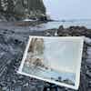 An open sketchbook sits on a snowy and rocky seashore, with a painting of the landscape visible on the sketchbook.