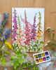A watercolor painting of foxgloves sits on a wooden table, with wildflowers and a watercolor palette.