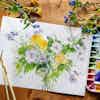 A watercolor painting of wildflowers sits on a wooden table, with wildflowers and a watercolor palette.