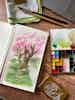 An open paint-filled palette and open notebook with a bright, cherry blossom sketch lay on a wooden table with various sketching supplies.