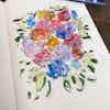 A sketchbook open to a ink and watercolor sketch of colorful flower buds, surrounded by leaves.
