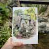 A hand holds up a sketchbook with a painting of a person sketching at the base of a small waterfall with a bridge.