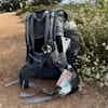 A grey-black backpack stuffed with supplies and art ziplocks pocking out sits on a dirt path lined with white wildflowers.