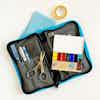 A Pocket Art Toolkit in grey with blue trim lays open on a white table, with an open Pocket Palette filled with colorful watercolors, a pair of scissors, a roll of tape, and a blue paper towel.