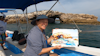 Maggie wears a sunhat and holds up a painting sketch of a rocky feature in the oceans while sitting on a boat.