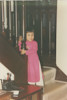 A small girl dressed in a long-sleeved pink dress smiles while gripping the bottom baluster of a banister inside.