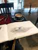 A café tabletop covered in sketching supplies, a partially-drawn mountain in a sketchbook, and a cappuccino. 